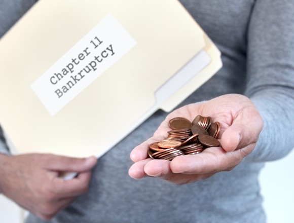 A man holds a manila folder marked Chapter 11 Bankruptcy, offering a handful of pennies in his other hand.