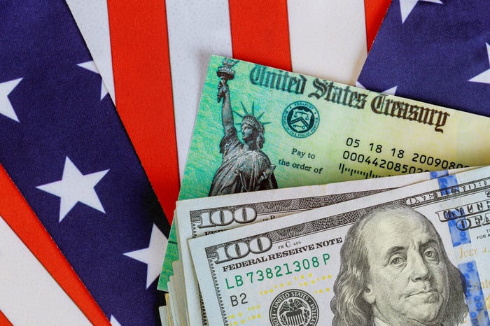 A stack of cash lying atop a U.S.Treasury check, which is surrounded by a folded American flag.