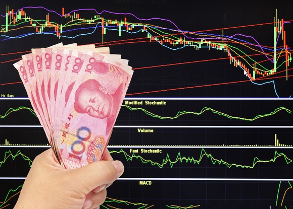 A hand holding yuan with stock charts in the background.