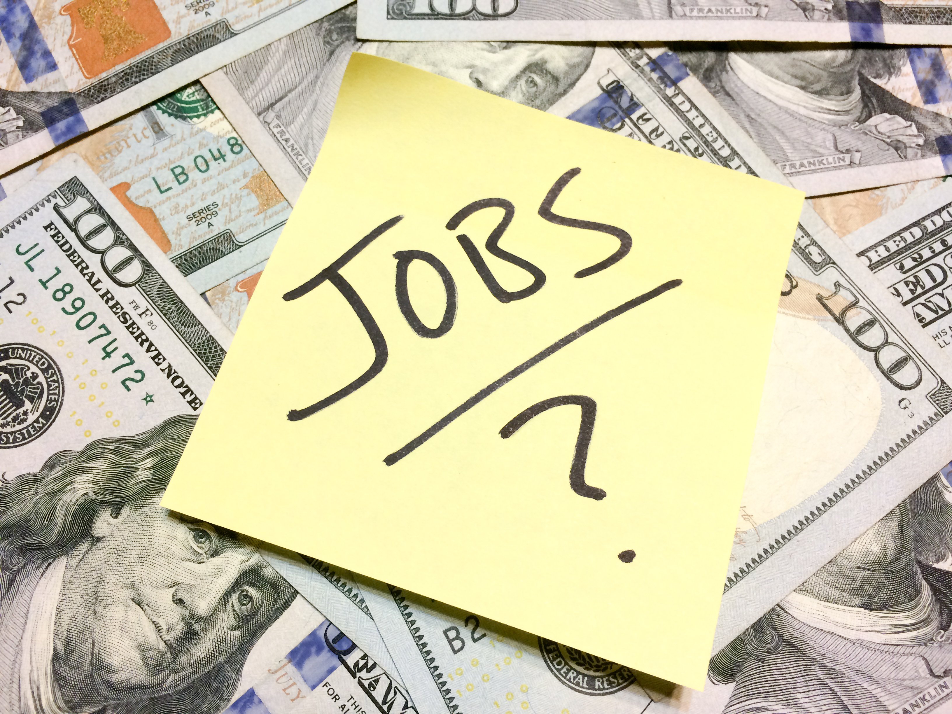 The word jobs with a question mark on a yellow sticky note on stop of hundred-dollar bills_GettyImages-1202347400