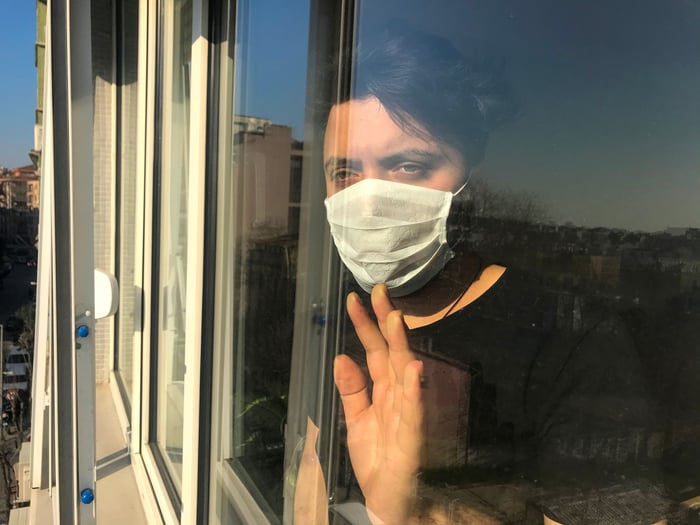 Sad woman in a face mask places hand on the window and looks outside