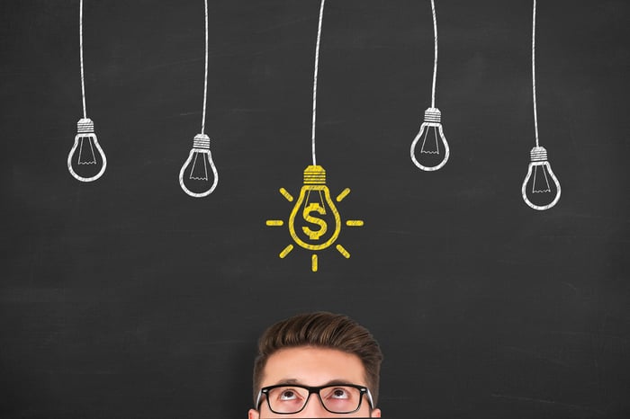 Man looking up at chalkboard drawings of light bulbs with one light bulb colored yellow with a dollar sign in it