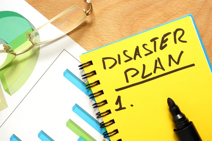 A yellow notepad with the words "disaster plan" written on it rests on a table.