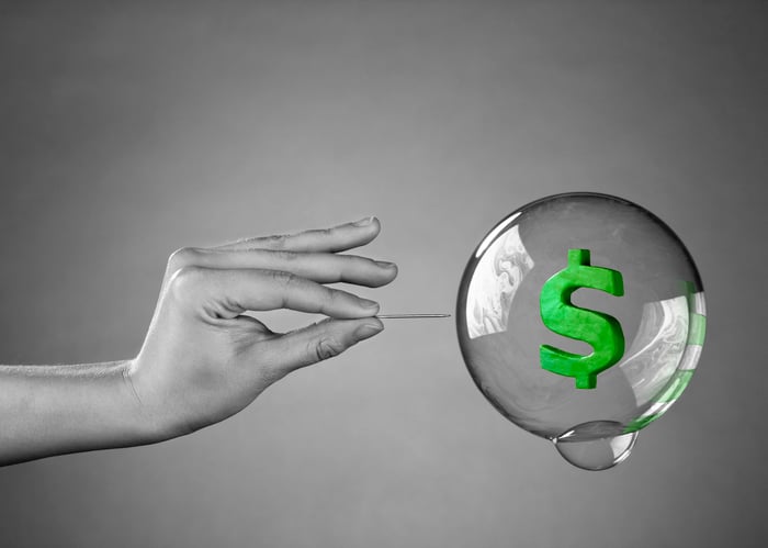 A grayscale hand holding a needle is poised to pop a bubble containing a green dollar sign.