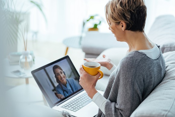 A woman sits on a white couch holding a mug and talking to another woman through a laptop video chat.