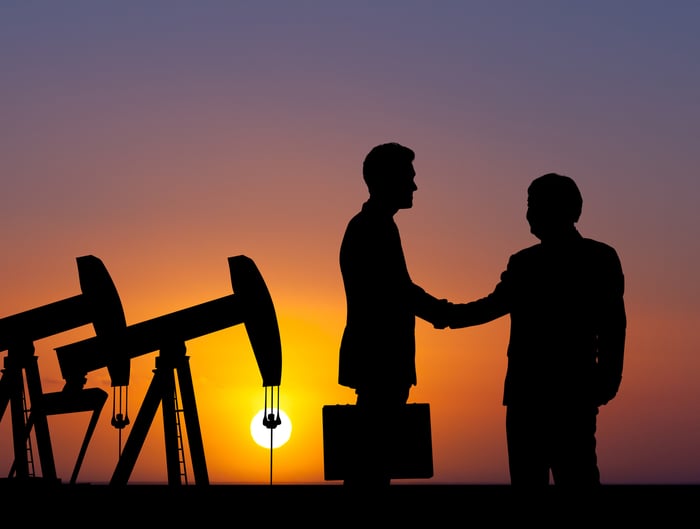 The silhouettes of two people shaking hands with oil pumps in the background.