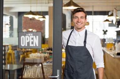 Smiling man in apron standing in restaurant doorway next to a sign stating that they're open_GettyImages-544353792