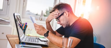 Man at laptop holding documents while holding his head_GettyImages-614422002
