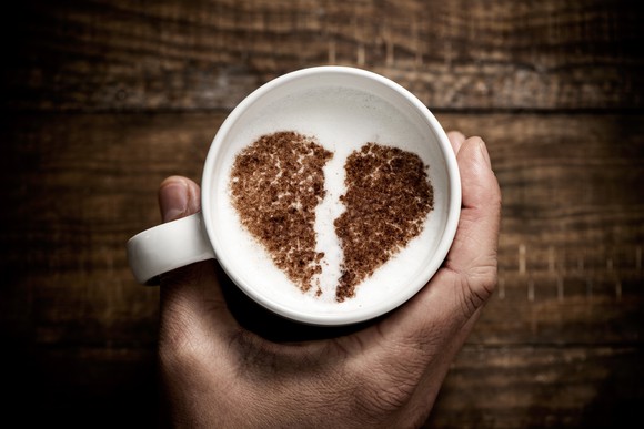 a white ceramic cup of cappuccino, with a broken heart drawn with cocoa powder on its milk foam