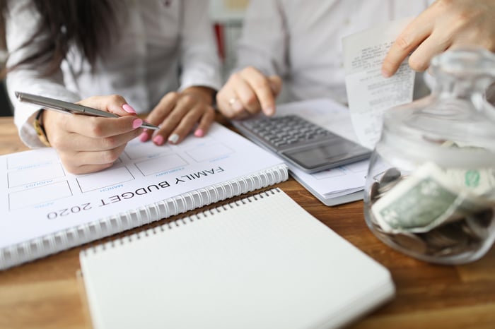 Two people creating a family budget at a table, with calculator and a jar with money on top of the table.