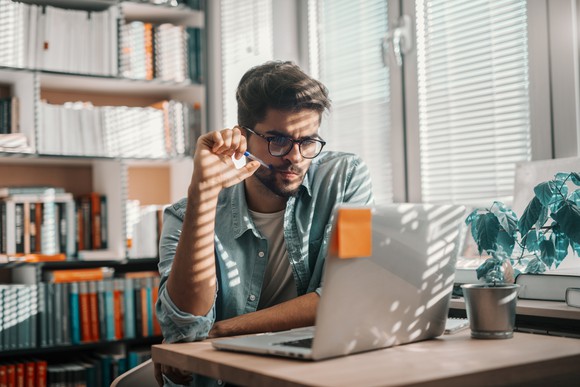 Man sitting at laptop with serious expression resting pen on face
