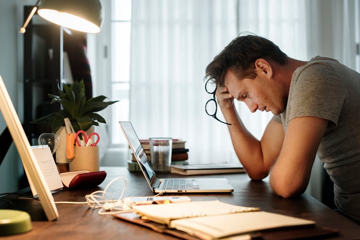 Man at laptop looking down with pained expression while holding eyeglasses