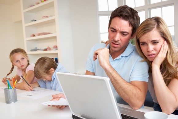 A visibly concerned couple reading material on a laptop, with their kids in the background.