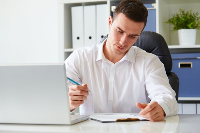 Man at table holding pen with laptop and clipboard in front of him