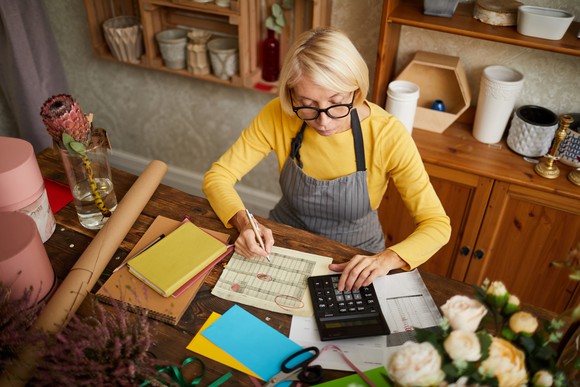 Woman in apron at table holding pen over document and typing on calculator