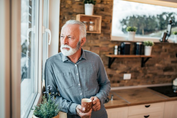 Older man with serious expression holding mug while looking out window