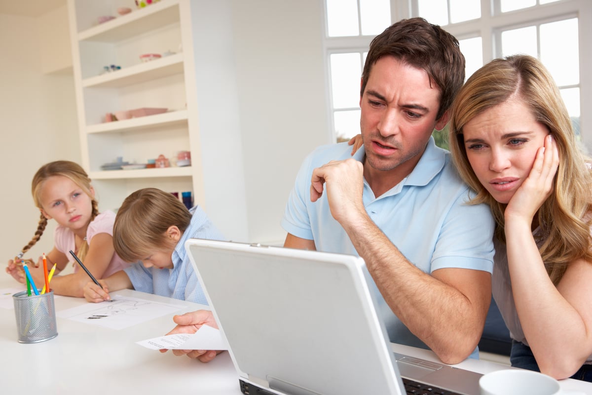 A visibly concerned couple looking at their laptop, with their kids in the background.