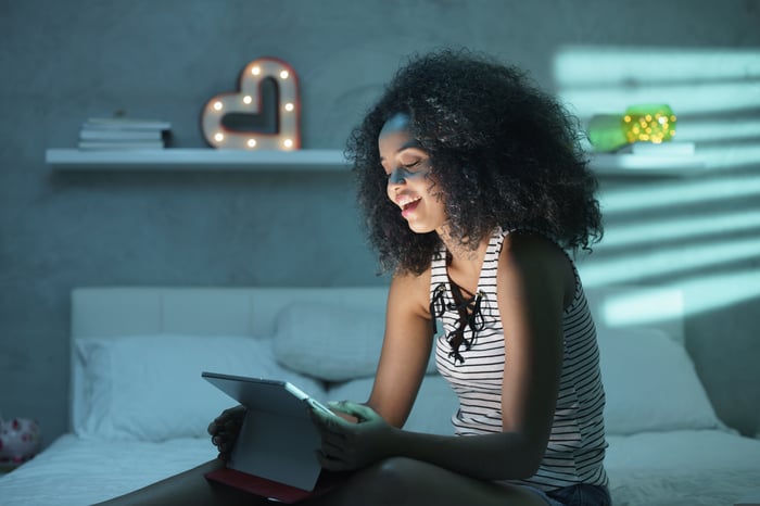 Smiling young women sitting on a bed watching streaming video on a tablet at night.