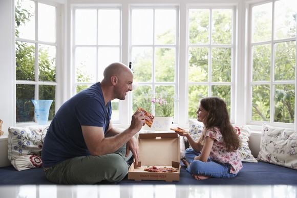 Father and daughter sitting on a window seat and eating pizza out of a box.