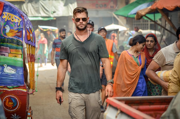 In a scene from a movie, a man wearing cargo pants, a t-shirt, and sunglasses walking through a colorful outdoor market in India.