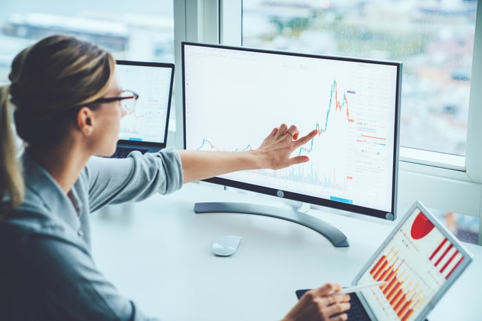 A woman working on a laptop and desktop simultaneously, and pointing to a chart.