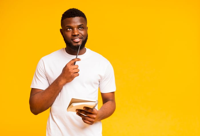 A young man thinks while holding a notepad against an orange background.