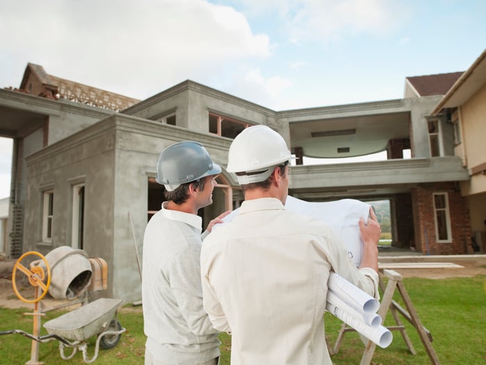 Construction workers examine blueprints in front of a house under construction.