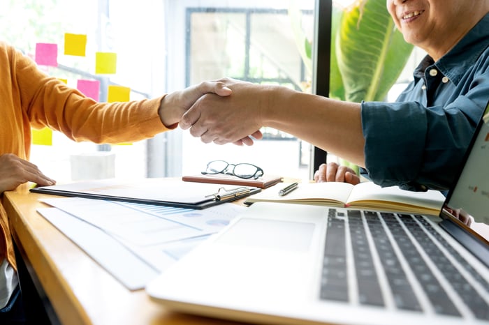 A smiling businessman shakes hands across a table with a woman in a yellow sweater, with an open laptop in the foreground