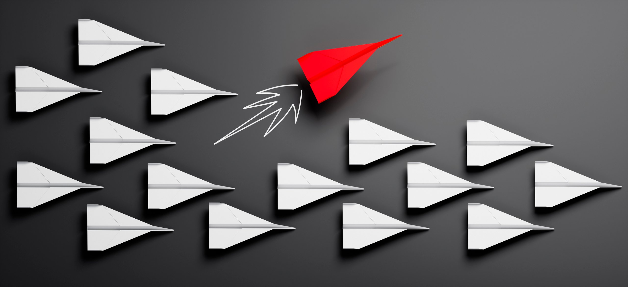 A red paper airplane flies in a different direction than a group of white paper airplanes.