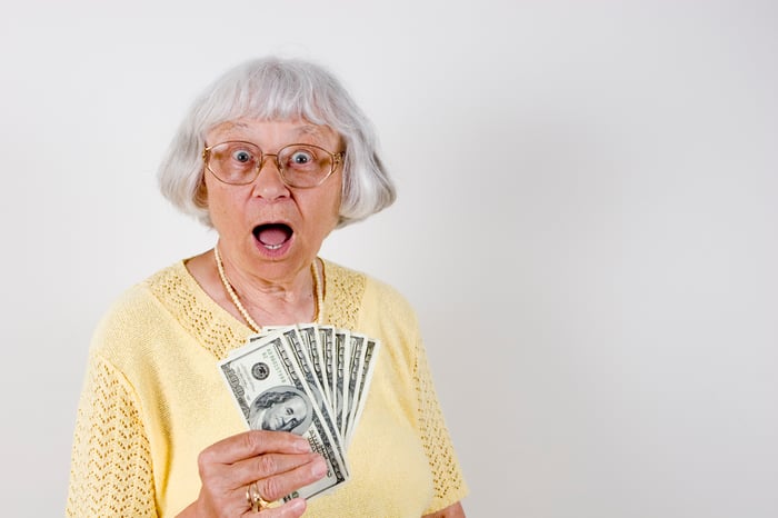 An older woman is holding a bunch of hundred dollar bills, with her mouth open in surprise.