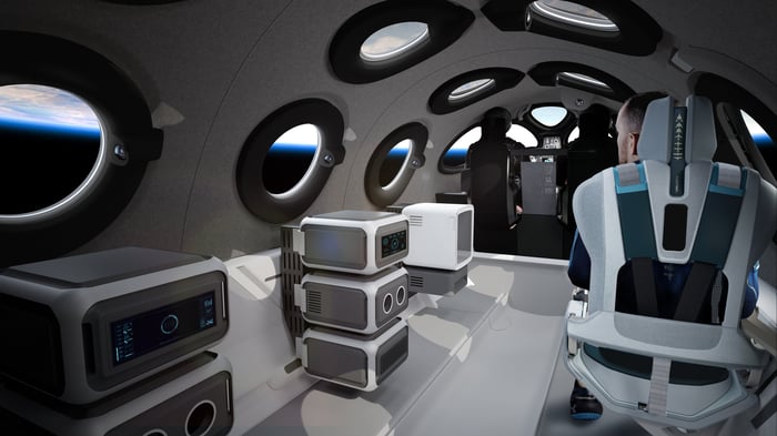 Interior photo of VSS Unity showing a passenger sitting next to some high-tech looking boxes.