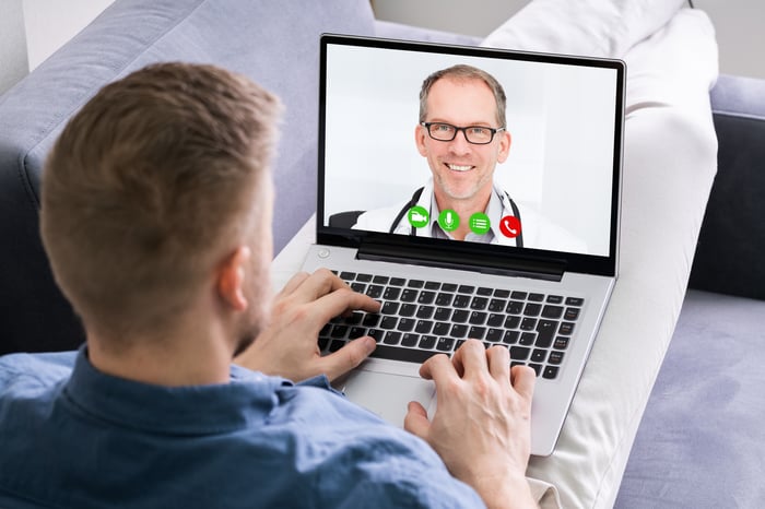 Young man leaning back on chair or sofa using a laptop that has a middle-aged male physician on screen.