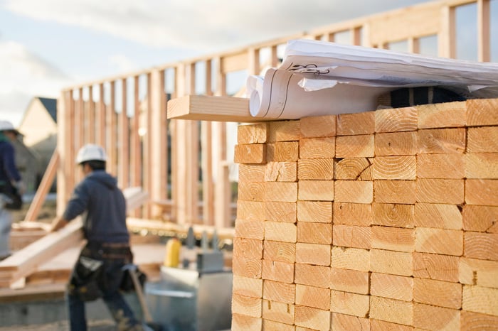 A stack of 2 by 4s at a construction site, with a worker in the background.