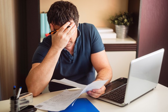 Man at laptop holding documents and covering his face