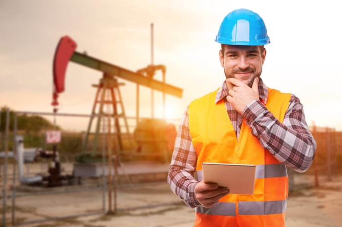 A worker standing in front of an oil rig with tablet in his hand