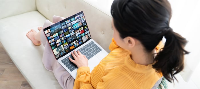 A woman with bare feet sitting on a couch looking at streaming video options on a laptop. 