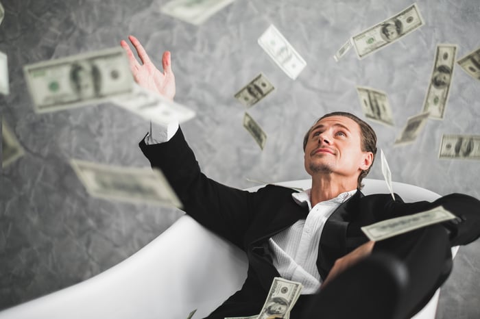 Man in a tuxedo sitting in a bathtub catching falling $100 bills. 