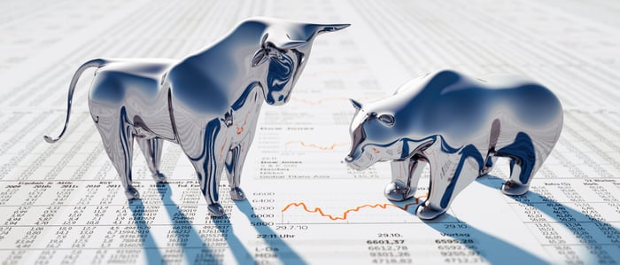 A silver bull standing tall next to a submissive silver bear, both of which are atop a newspaper with stock quotes.