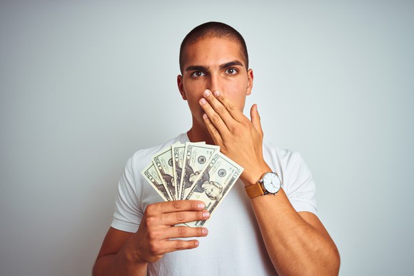 Man covering his mouth in embarrassment while holding five $20 bills.