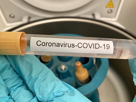 Gloved hand holding test tube with "Coronavirus-COVID-19" printed on label and a centrifuge in the background