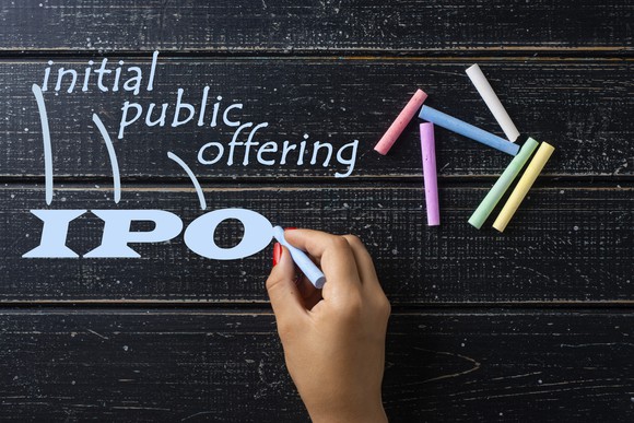 A hand draws “IPO” with a piece of blue chalk.