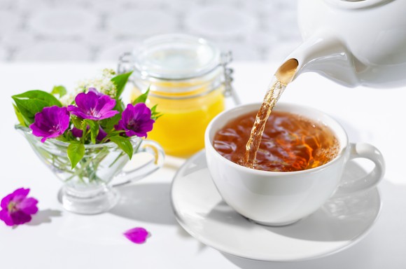 A cup of tea being poured from a teapot next to some flowers in a crystal teacup