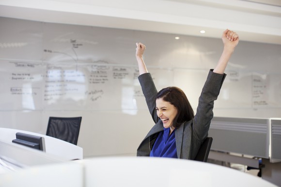 Person at desk with arms raised high and big smile, with whiteboard behind.