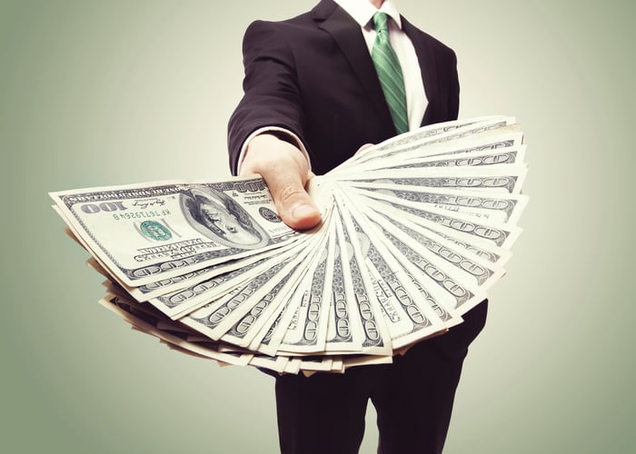 A person in a business suit is holding out $100 bills.