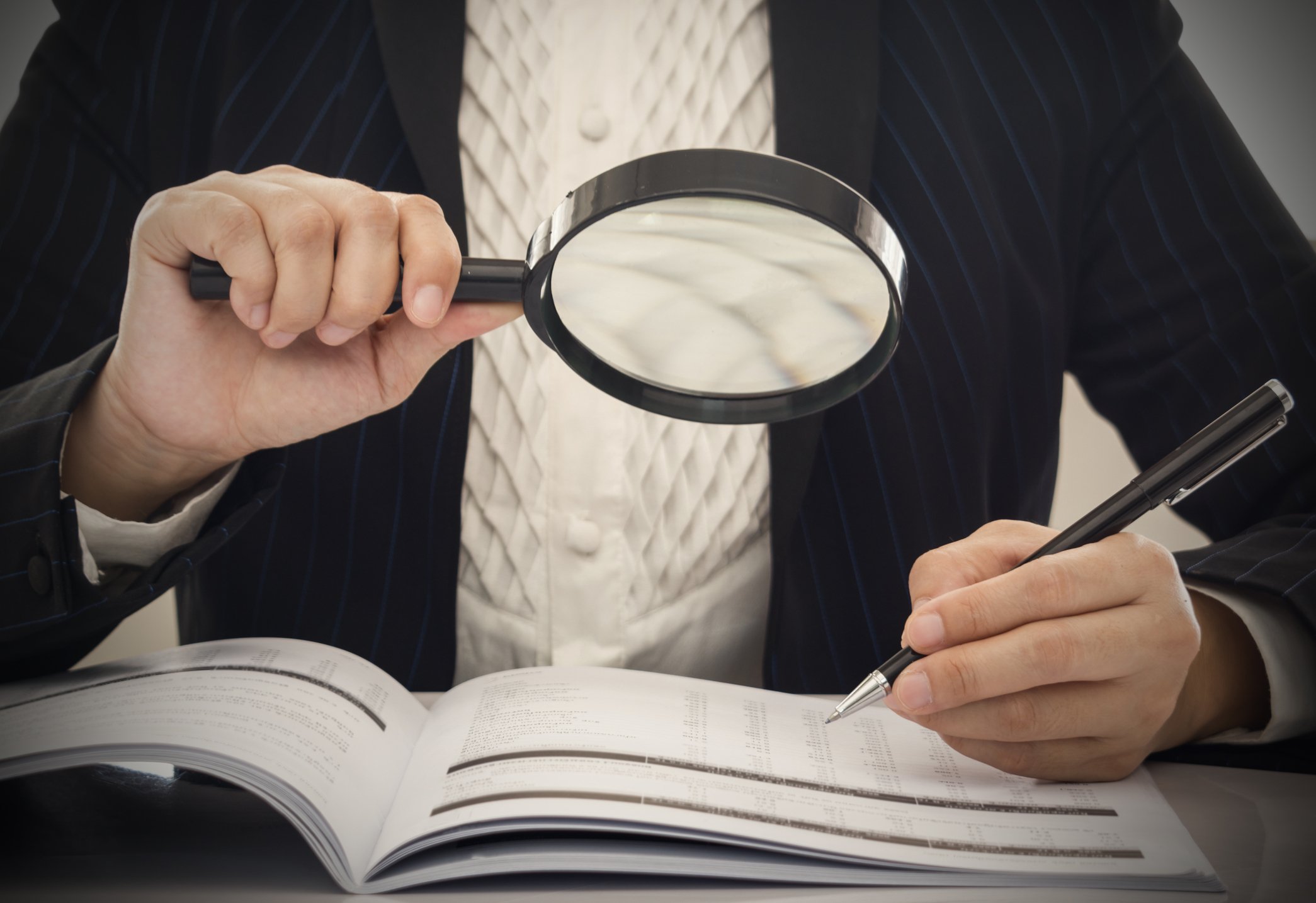 Auditor in business suit examines financial records with a pen and a magnifying glass