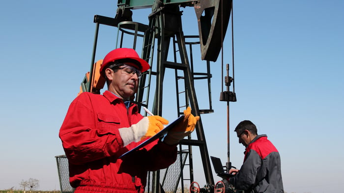 An oil well and two workers, one worker in the foreground writing in a notebook and the other in the background writing on a laptop. 