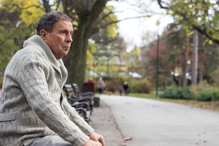 Older man in sweater with serious expression, sitting on park bench.