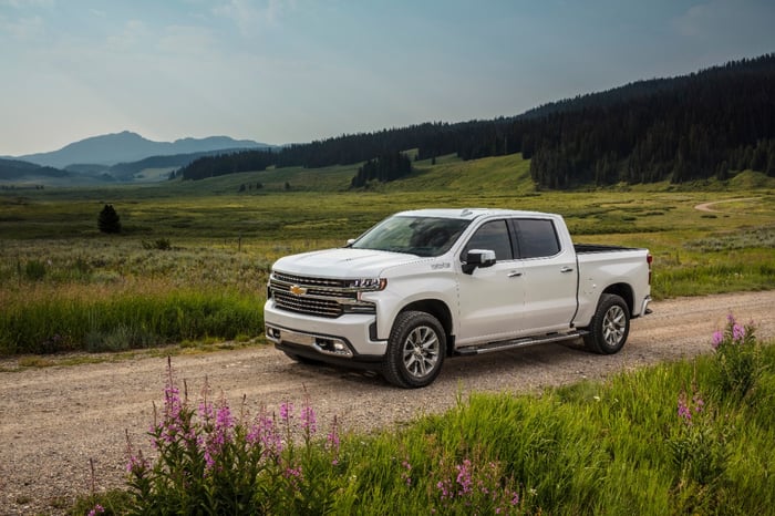 A white Chevy Silverado driving on a dirt road, with a green field in the background