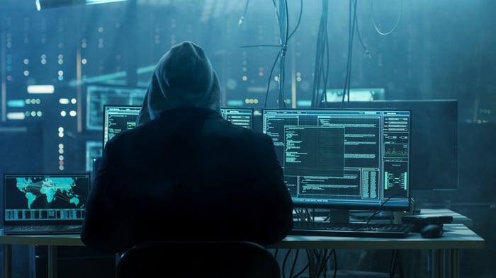 A hooded hacker in a darkened room full of computers, monitors, and cables.