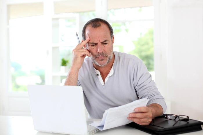 Man who seems confused looking at a some papers while sitting in front of a computer.